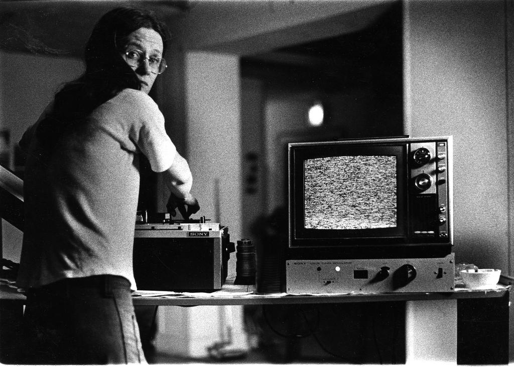 black and white photograph. a woman with long hair and glasses looks over her shoulder while she works with what could be image editing equipment sitting next to a television should static on its screen