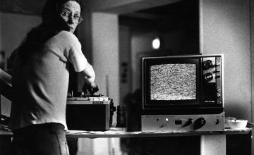 black and white photograph. a woman with long hair and glasses looks over her shoulder while she works with what could be image editing equipment sitting next to a television should static on its screen