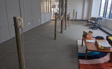 long, rectangular room with grey carpet and beige walls. room is bisected lengthwise by a barbed wire fence. a yellowing phone from the 1980s is affixed to one wood post. at the opposite end of the room a black Bell telephone from 1915 or so is affixed to another post. to the right of the fence is a colorful rainbow shag carpet with a coffee table and couch.