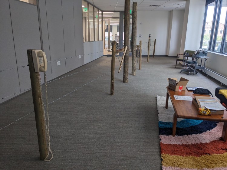 long, rectangular room with grey carpet and beige walls. room is bisected lengthwise by a barbed wire fence. a yellowing phone from the 1980s is affixed to one wood post. at the opposite end of the room a black Bell telephone from 1915 or so is affixed to another post. to the right of the fence is a colorful rainbow shag carpet with a coffee table and couch.