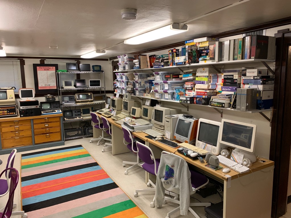 room in the basement of a house. rainbow carpet. purple chairs placed in front of fake wood metal desks on top of which are desktop computers from the early 1980s to early 2000s. Above the computers are two shelves full of boxes of software stacked on top of each other. in the background is a metal shelf with portable computers and laptops.